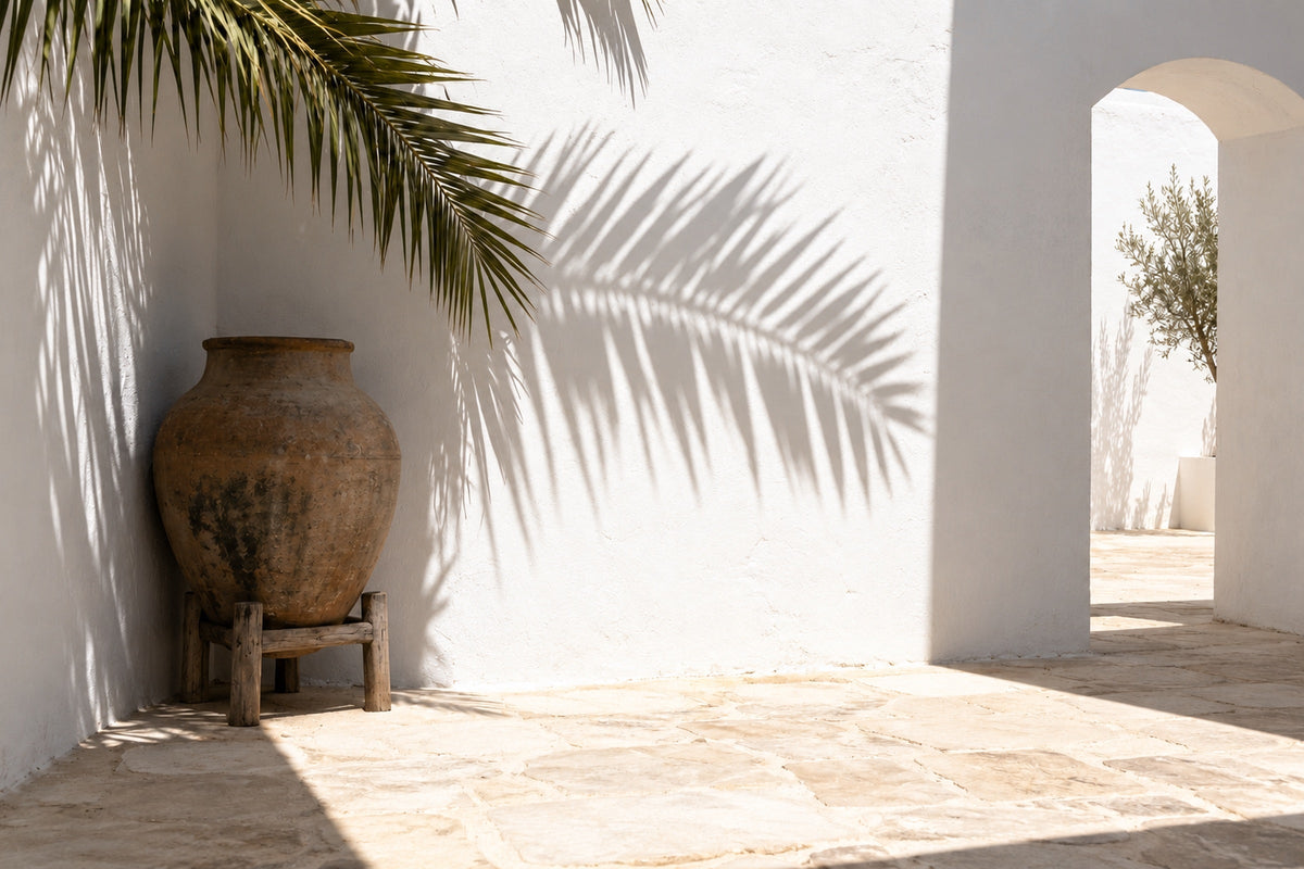 A white courtyard at midday in summer, with a clay water jar on a wooden stand in the corner and the shadow of a palm frond on the wall