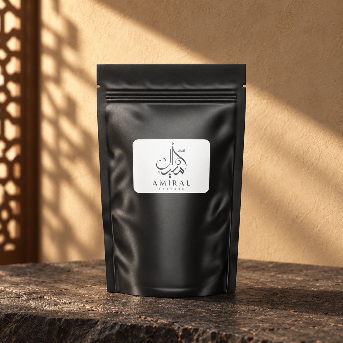 Amiral Bakhoor by Weight from Sedr Al-Khaleej; closed matte black pouch with a white label on basalt against a sand plaster wall, with angled sunlight and a wooden screen shadow.