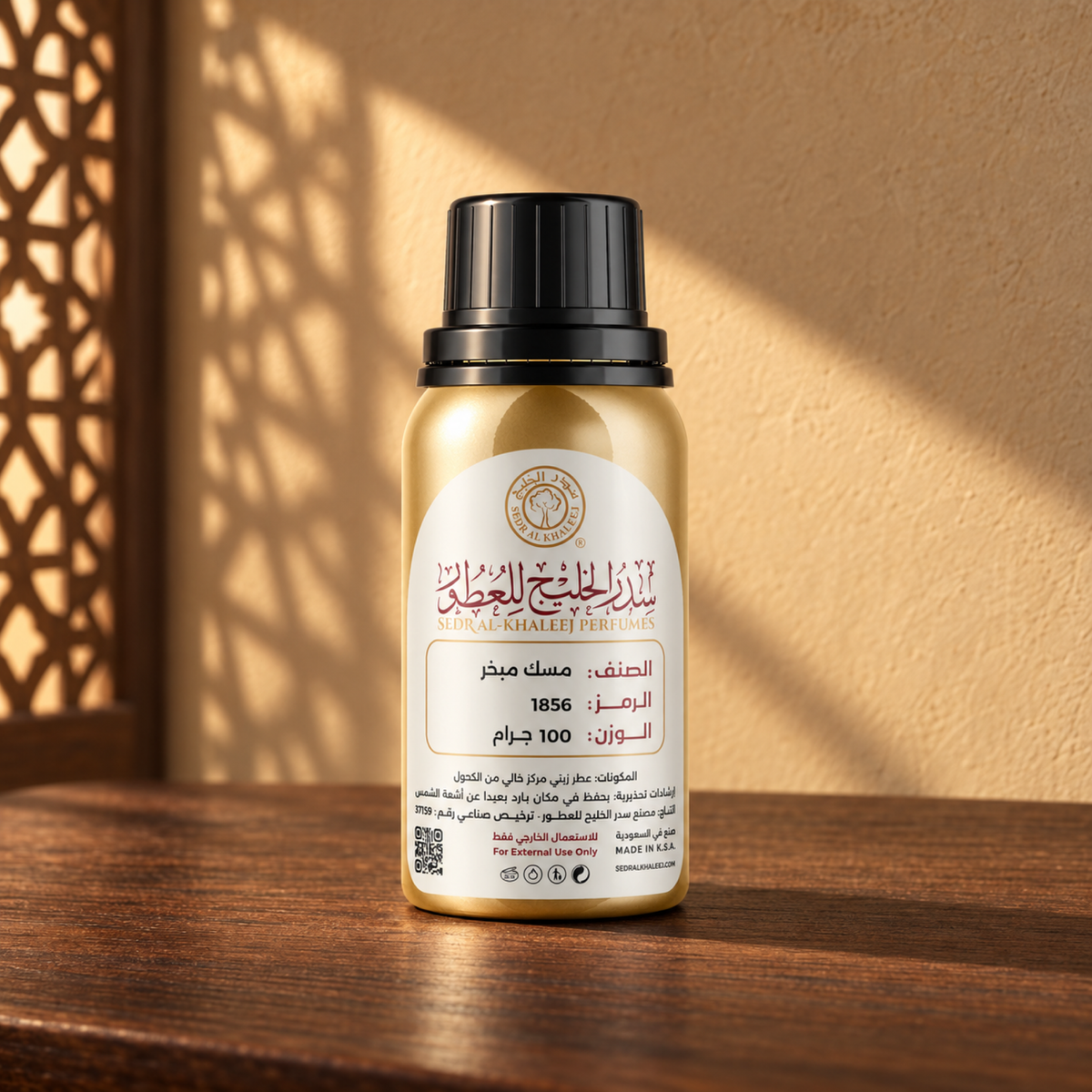 عبوة عطر مسك مبخر الزيتي من سدر الخليج، ذهبية بغطاء أسود وملصق كريمي، على خشب جوز أمام جدار رملي مضاء بالشمس وظل مشربية.