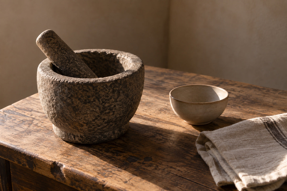 A stone mortar and pestle on an old wooden table, beside a small ceramic dish and a cloth napkin