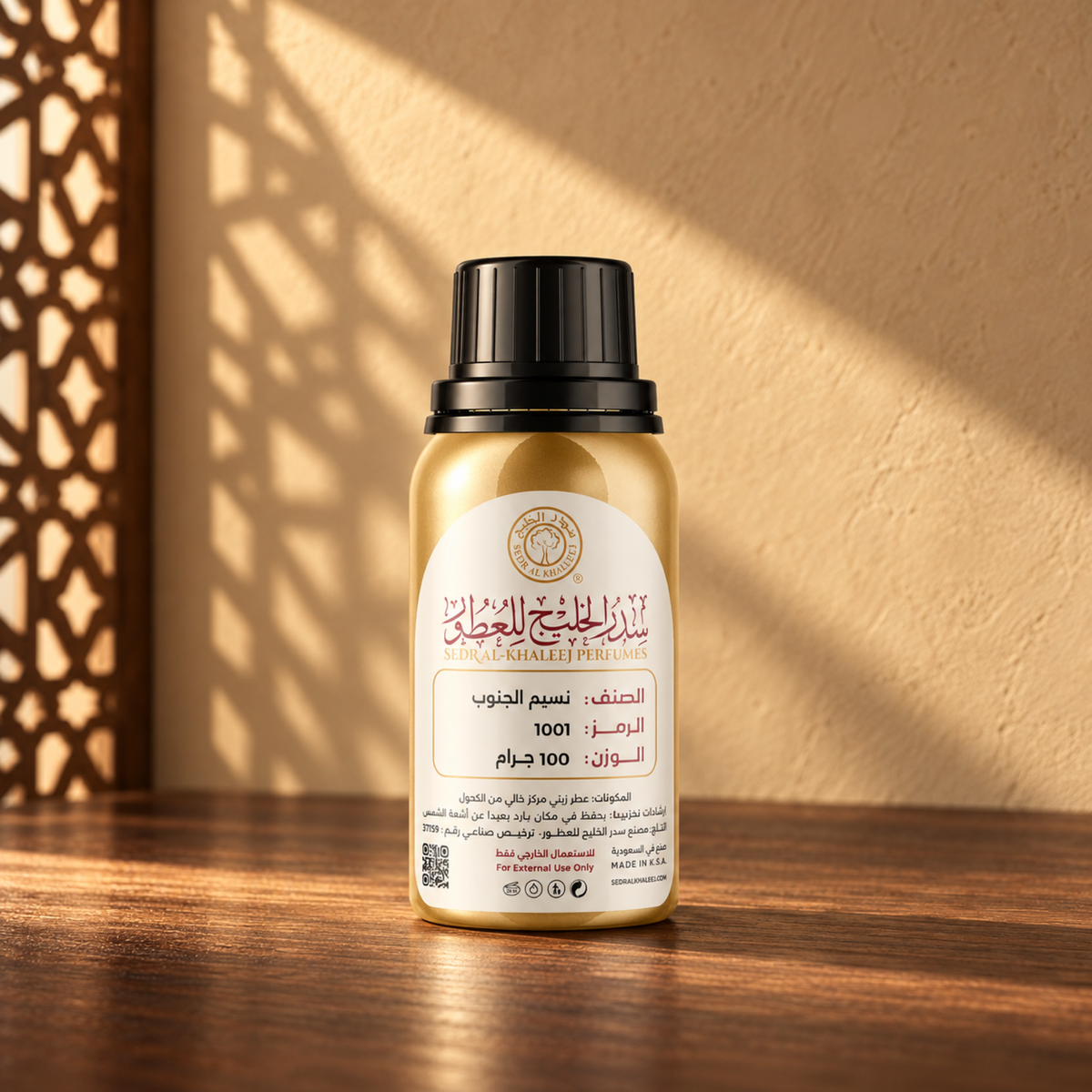 Sedr Al-Khaleej Naseem Aljanob perfume oil in a closed gold bottle with a black cap and cream label on walnut wood against a sand wall in sunlight beside a screen shadow.