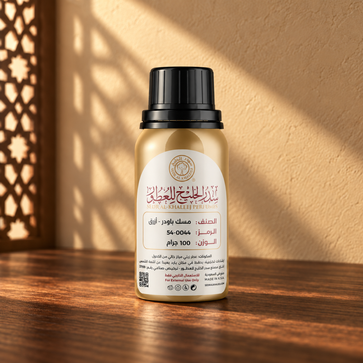 Sedr Al-Khaleej Blue Powder Musk perfume oil in a closed gold bottle with a black cap and cream label on walnut wood against a sand wall in sunlight beside a screen shadow.