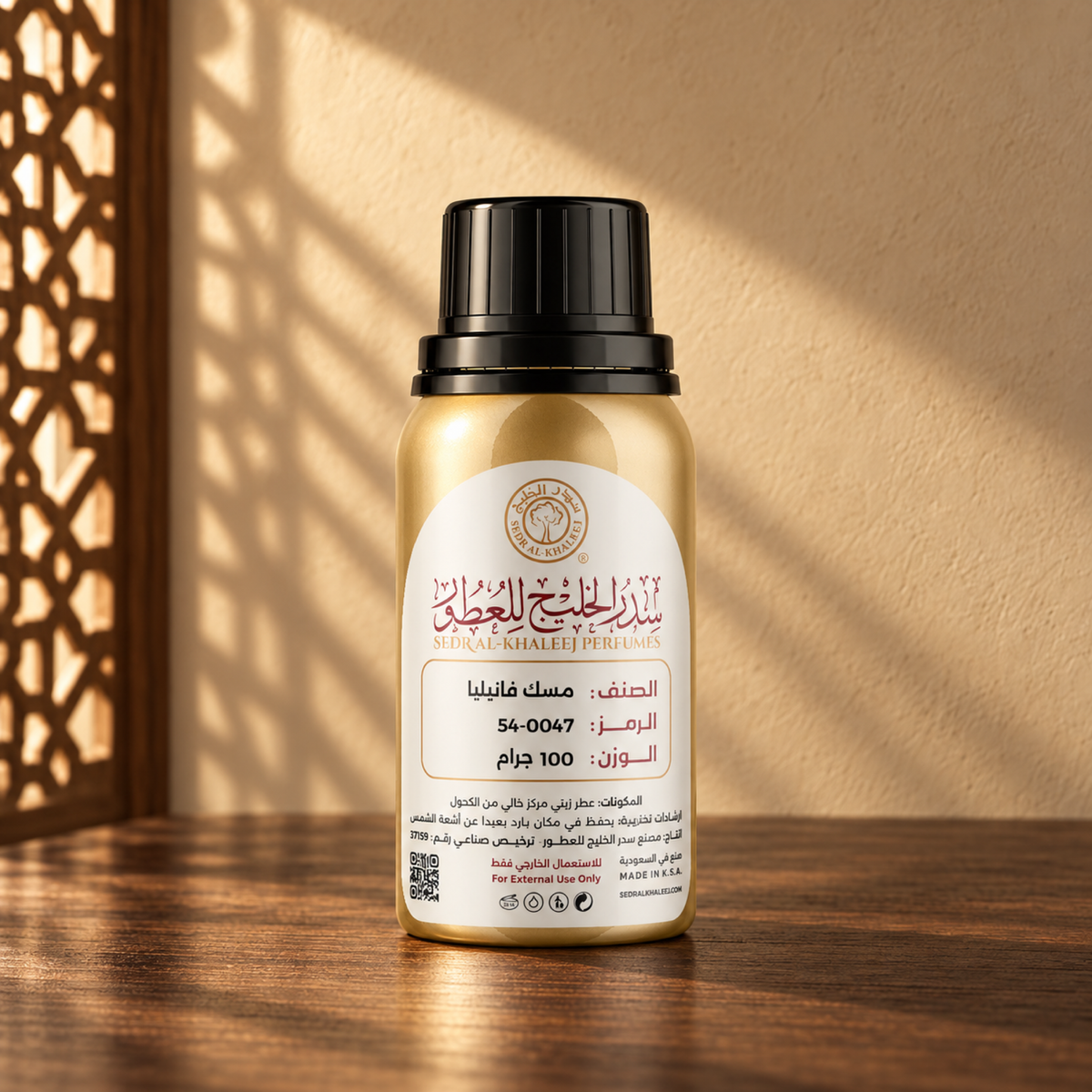 عبوة العطر الزيتي مسك فانيليا من سدر الخليج، ذهبية بغطاء أسود وملصق كريمي، على خشب جوز أمام جدار رملي في ضوء الشمس وظل مشربية.
