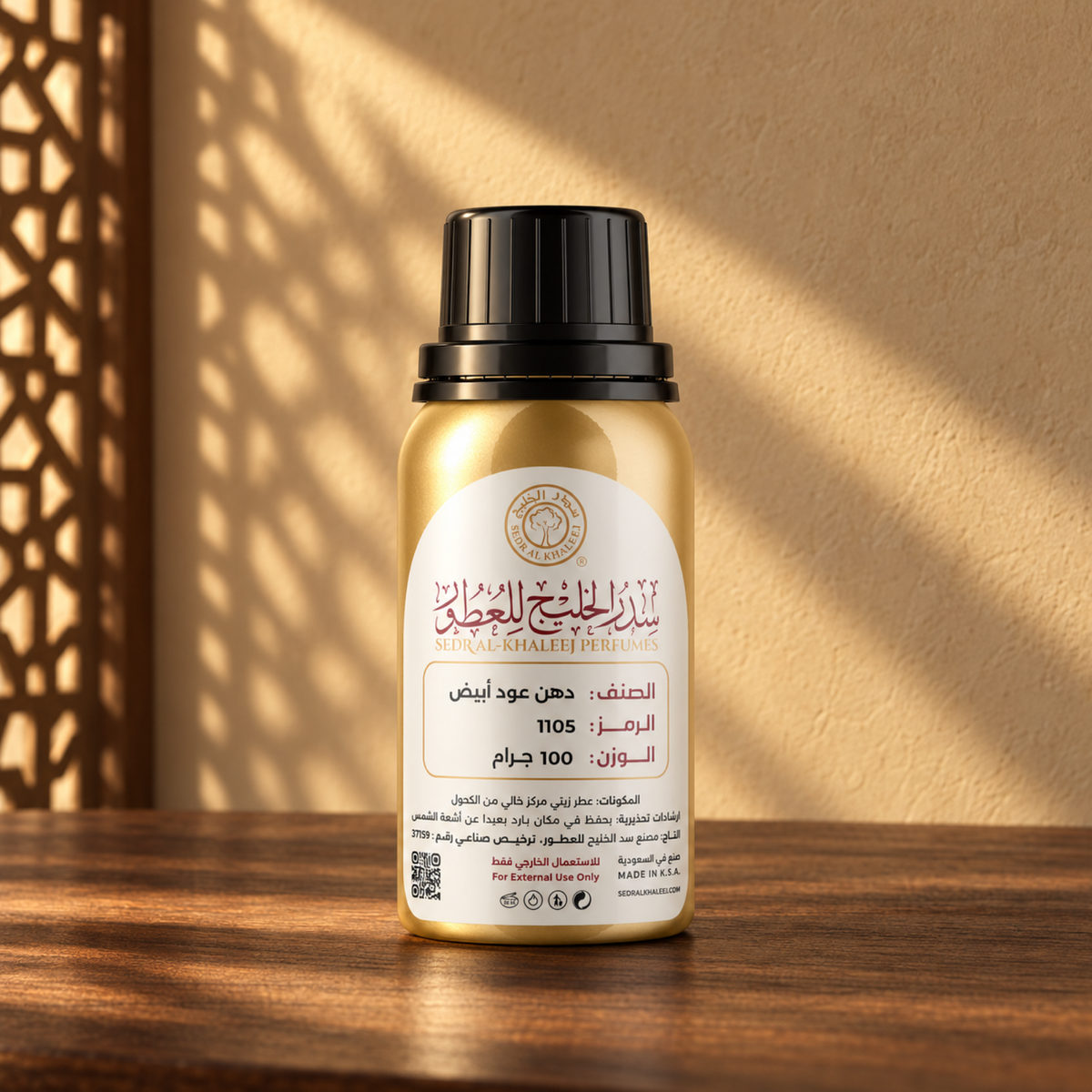 عبوة العطر الزيتي دهن عود أبيض من سدر الخليج، ذهبية بغطاء أسود وملصق كريمي، على خشب جوز أمام جدار رملي في ضوء الشمس وظل مشربية.